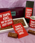 Crude Crumbs Christmas gift set featuring a brownie box, brownie slices, red Christmas postcards, a matching coffee bag and mug, all displaying the festive quote “Santa loves a little sass.”