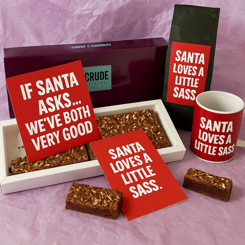 Crude Crumbs Christmas gift set featuring a brownie box, brownie slices, red Christmas postcards, a matching coffee bag and mug, all displaying the festive quote “Santa loves a little sass.”