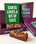 Purple Crude Crumbs brownie gift box opened to reveal a chocolate brownie slab topped with flakes, with a green postcard propped inside reading ‘Santa Loves a Bit of Sass’ in white Zuume Rough font.