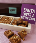 Christmas brownie gift box with Crude Crumbs branding, topped with chocolate curls, shown with a purple postcard reading “Santa loves a little sass” placed beside the open box.