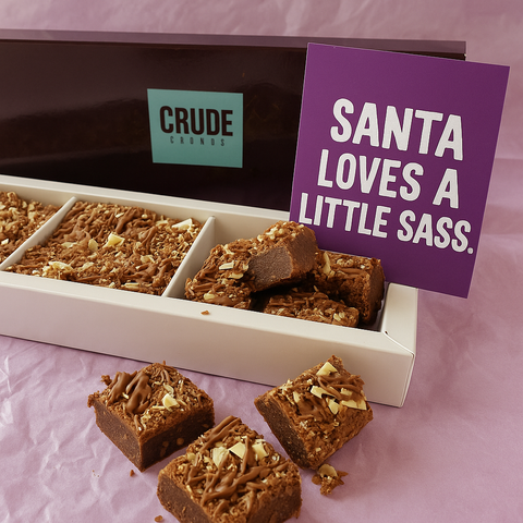 Christmas brownie gift box with Crude Crumbs branding, topped with chocolate curls, shown with a purple postcard reading “Santa loves a little sass” placed beside the open box.