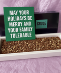 Large Crude Crumbs postal brownie slab in an open gift box, displayed with a green Christmas postcard reading “May your holidays be merry and your family tolerable” propped against the branded box.