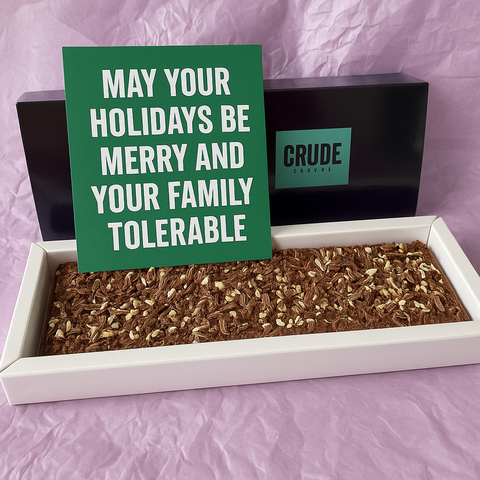 Large Crude Crumbs postal brownie slab in an open gift box, displayed with a green Christmas postcard reading “May your holidays be merry and your family tolerable” propped against the branded box.